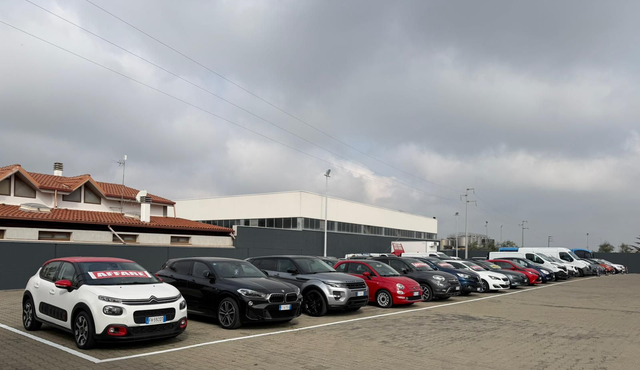Una fila di auto parcheggiate in un piazzale accanto alle case, sotto un cielo nuvoloso.