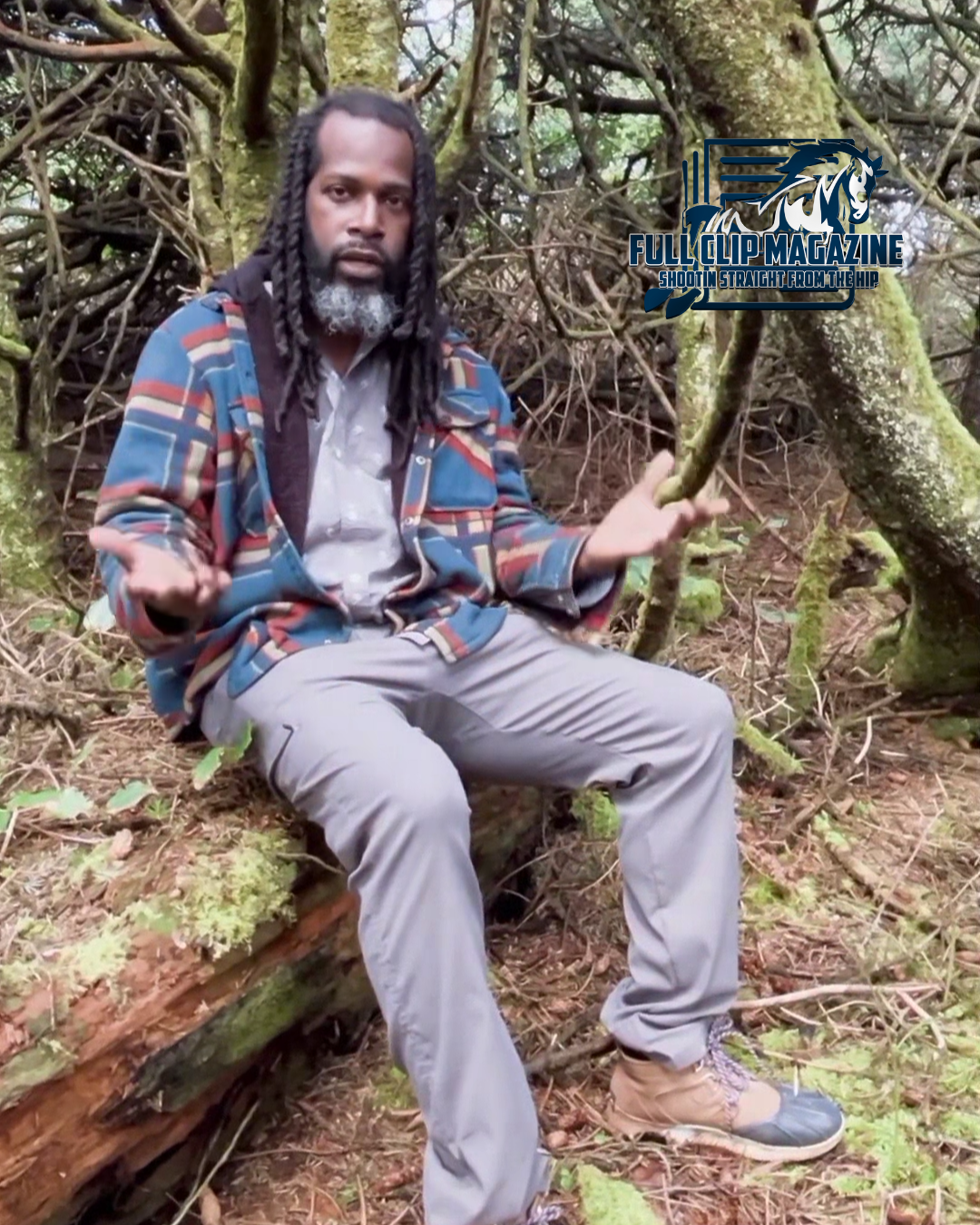 A man with dreadlocks is sitting on a log in the woods.