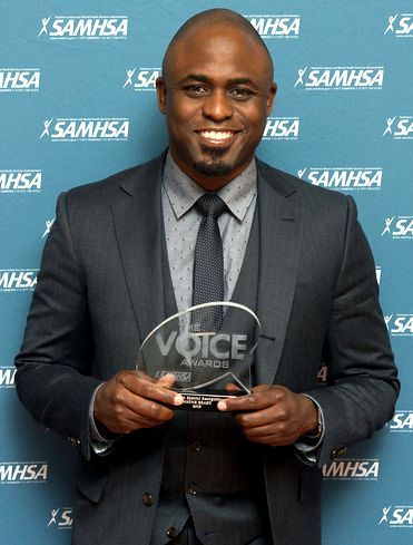 Man in suit holding an award, smiling in front of a backdrop with the SAMHSA logo.