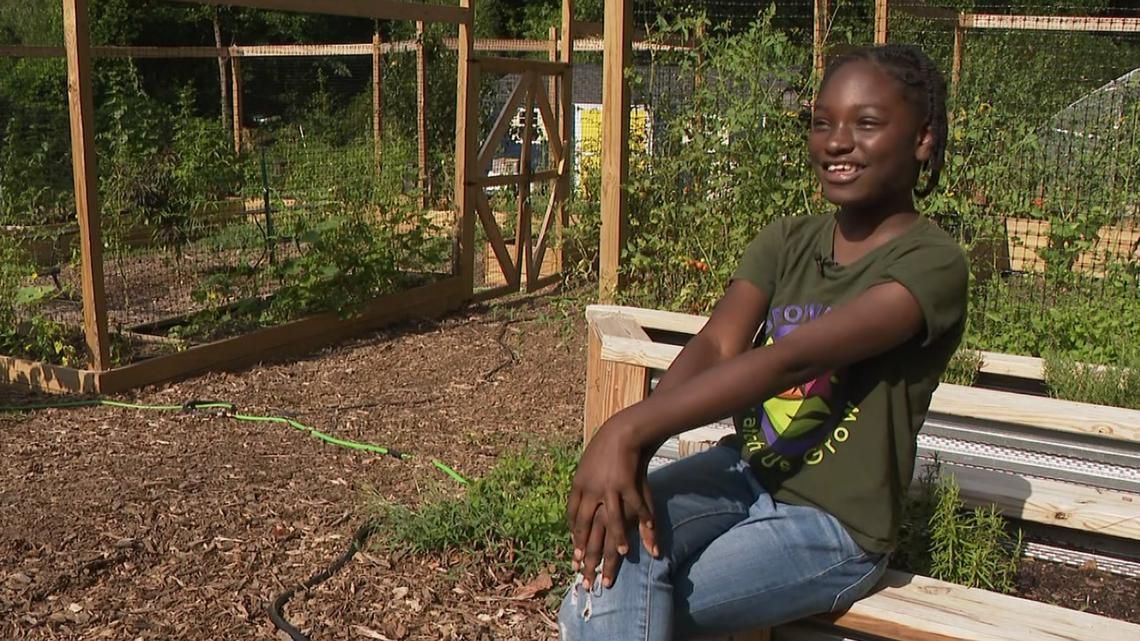 Young Black woman smiles, seated in a garden. Wooden structures and plants fill the background.