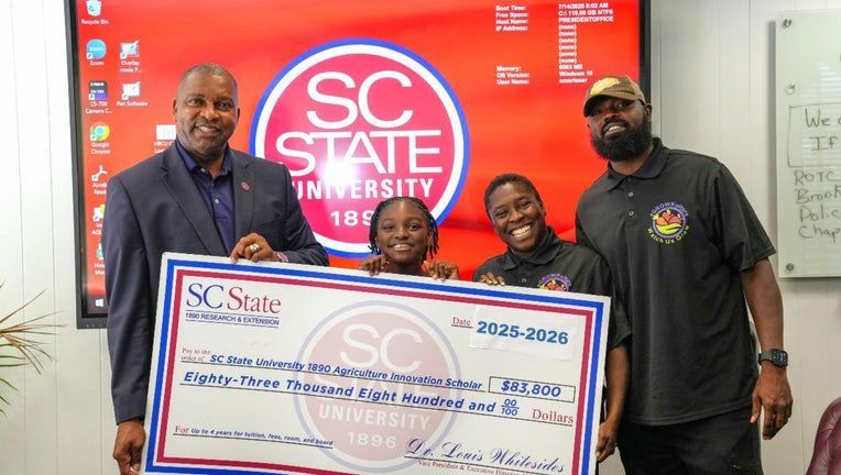 Four people holding a giant check for SC State University. Smiling in a room with a logo background.
