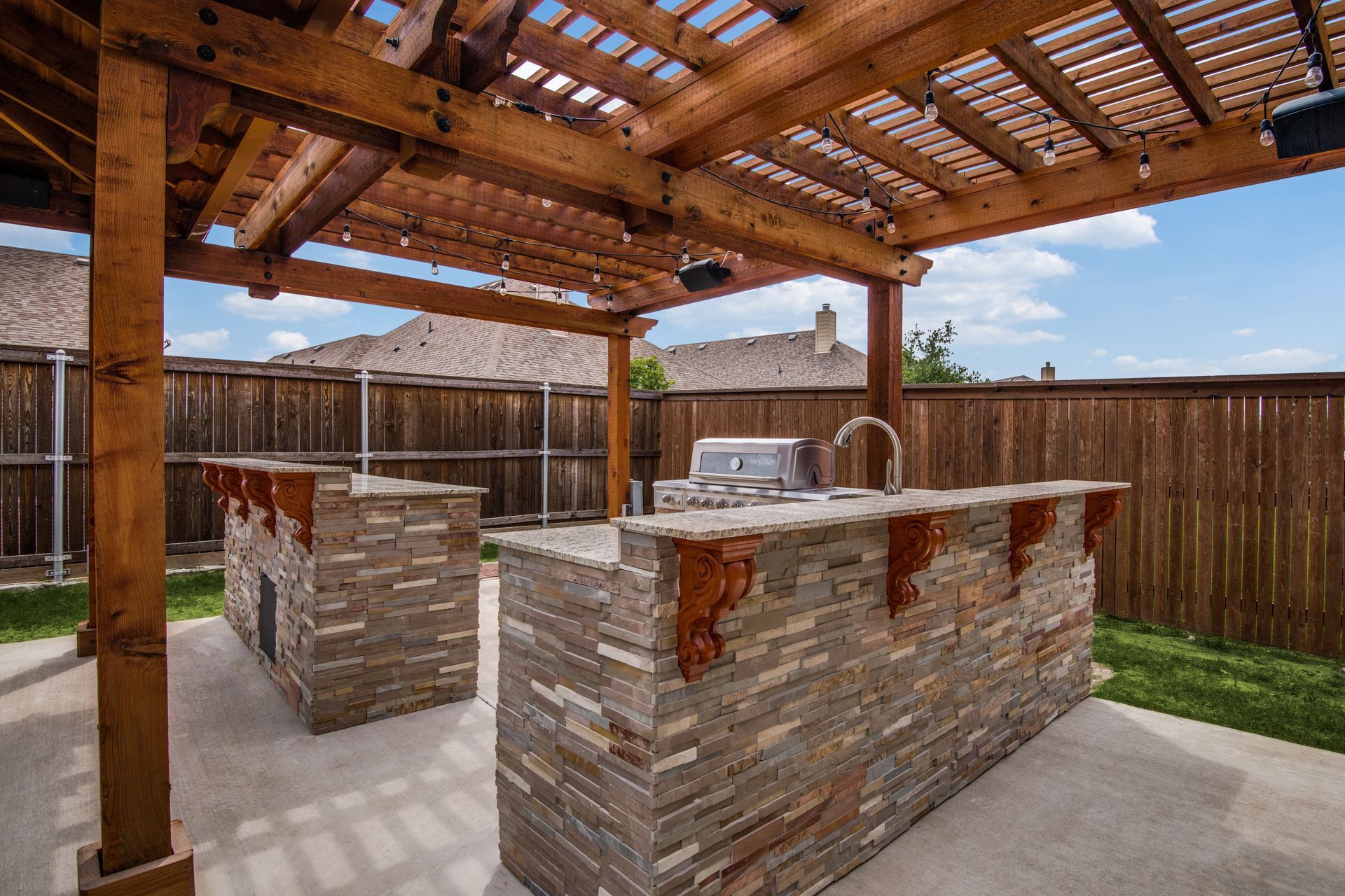 A patio with a wooden pergola and a stone island with a grill underneath it.