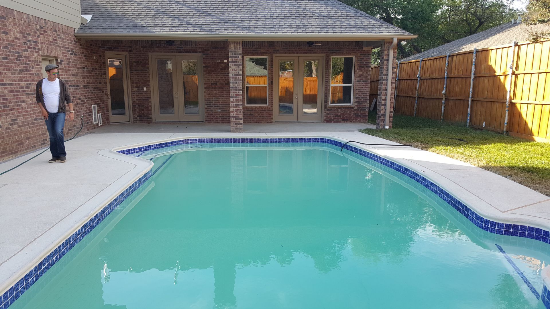 A man is standing next to a large swimming pool in front of a house remodel.