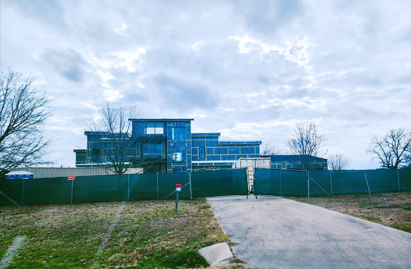 A large blue resilient design building is under construction behind a green fence.