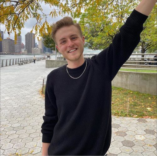 A young man in a black sweater is standing on a sidewalk with his arm in the air.