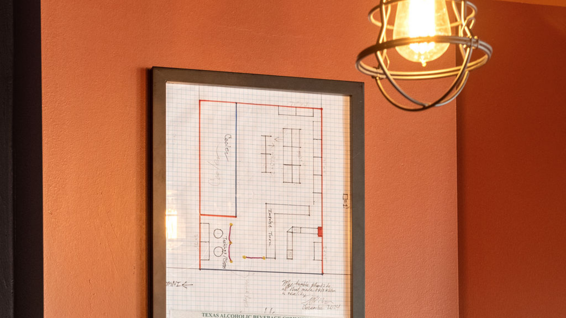 Framed blueprint on orange wall, lit by caged Edison bulb.