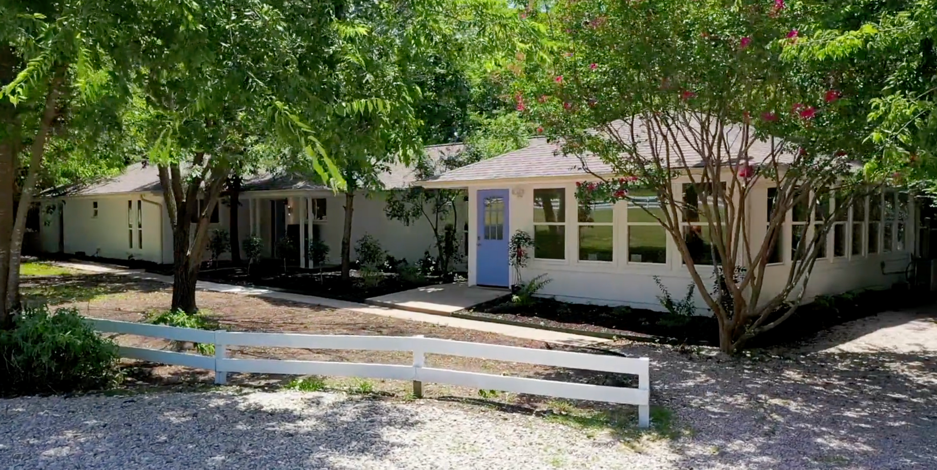 White house with blue door, white fence, and lush green trees in the yard.