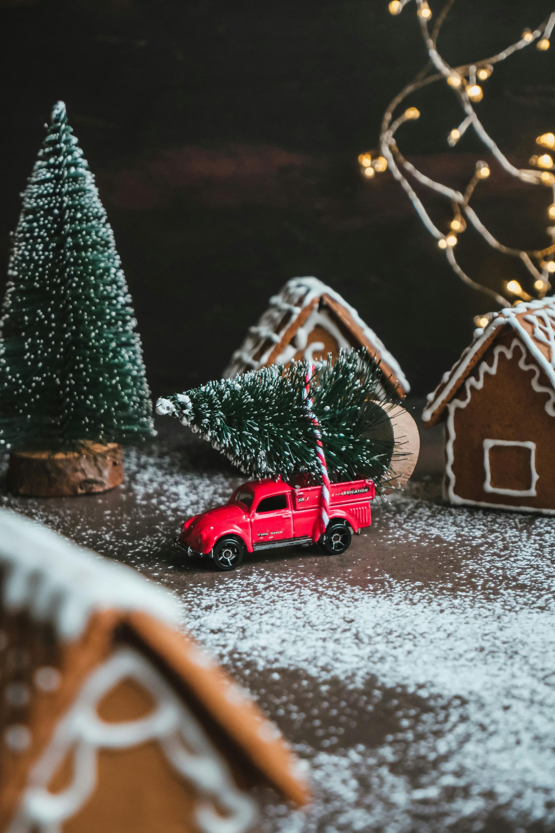 A red toy truck is carrying a christmas tree in a gingerbread village.