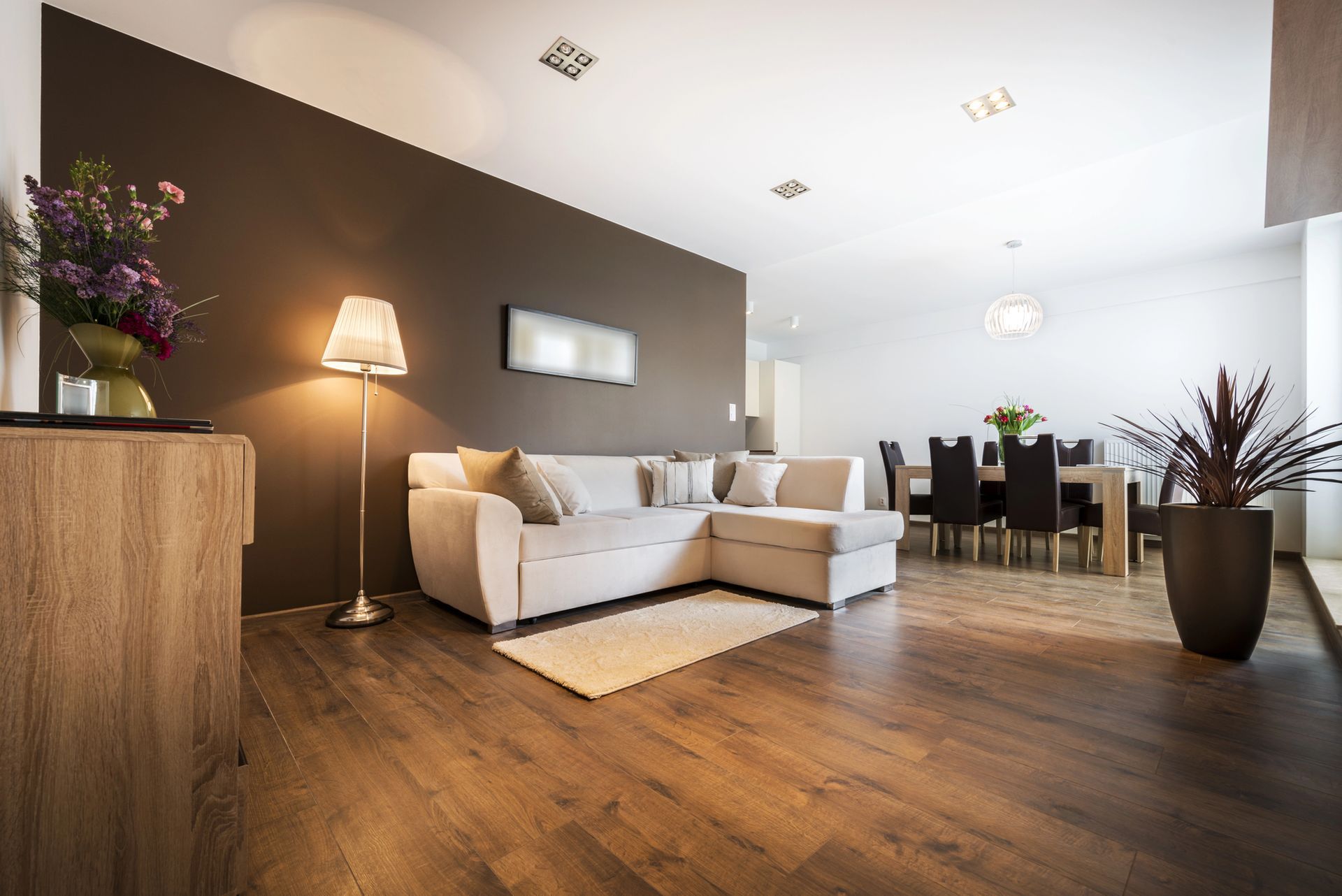 Salón moderno de planta abierta con sofá seccional blanco, suelos de madera, lámpara y zona de comedor.