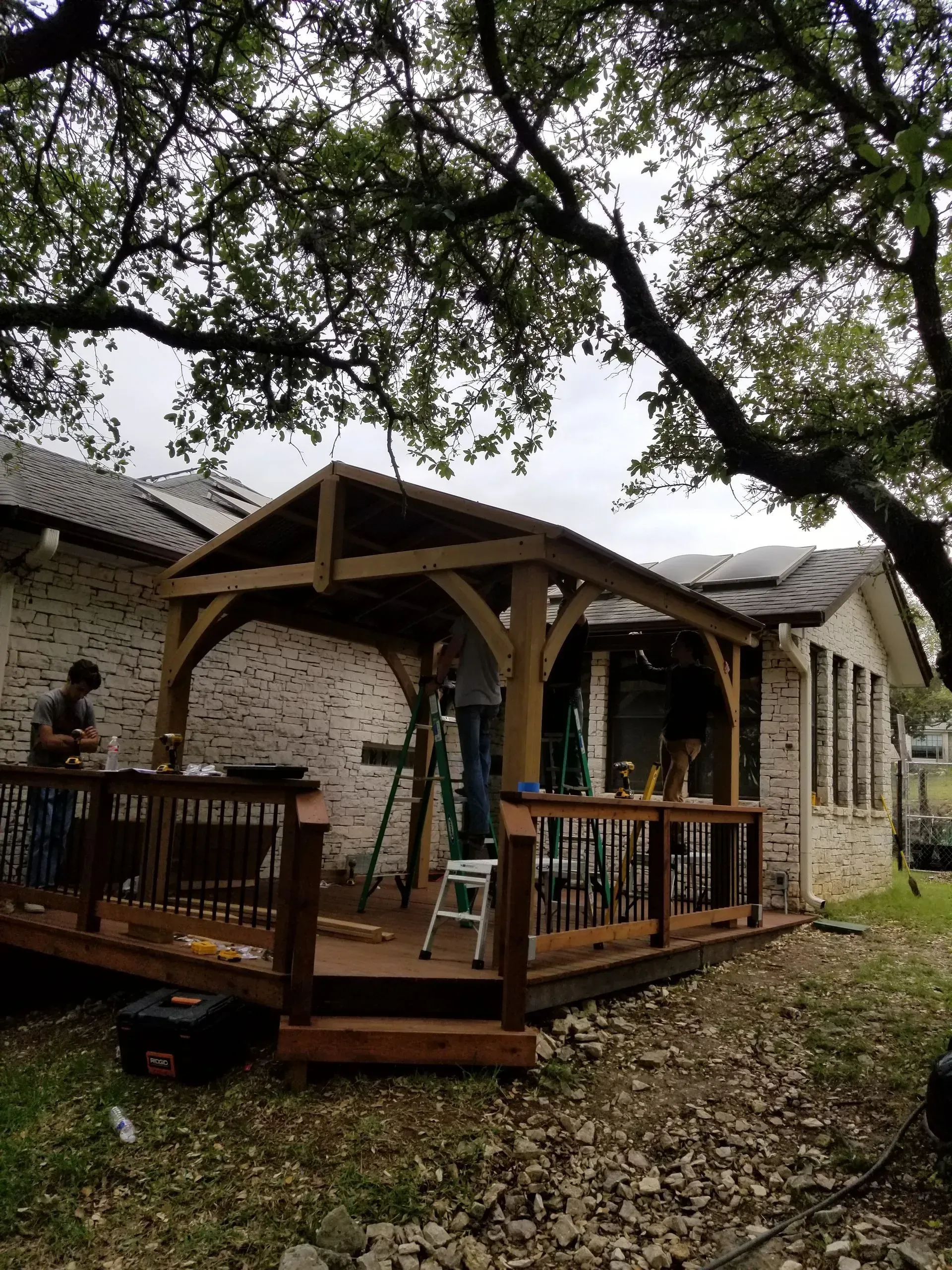 Wooden deck and arbor construction. Two workers on deck, building near brick house under tree.
