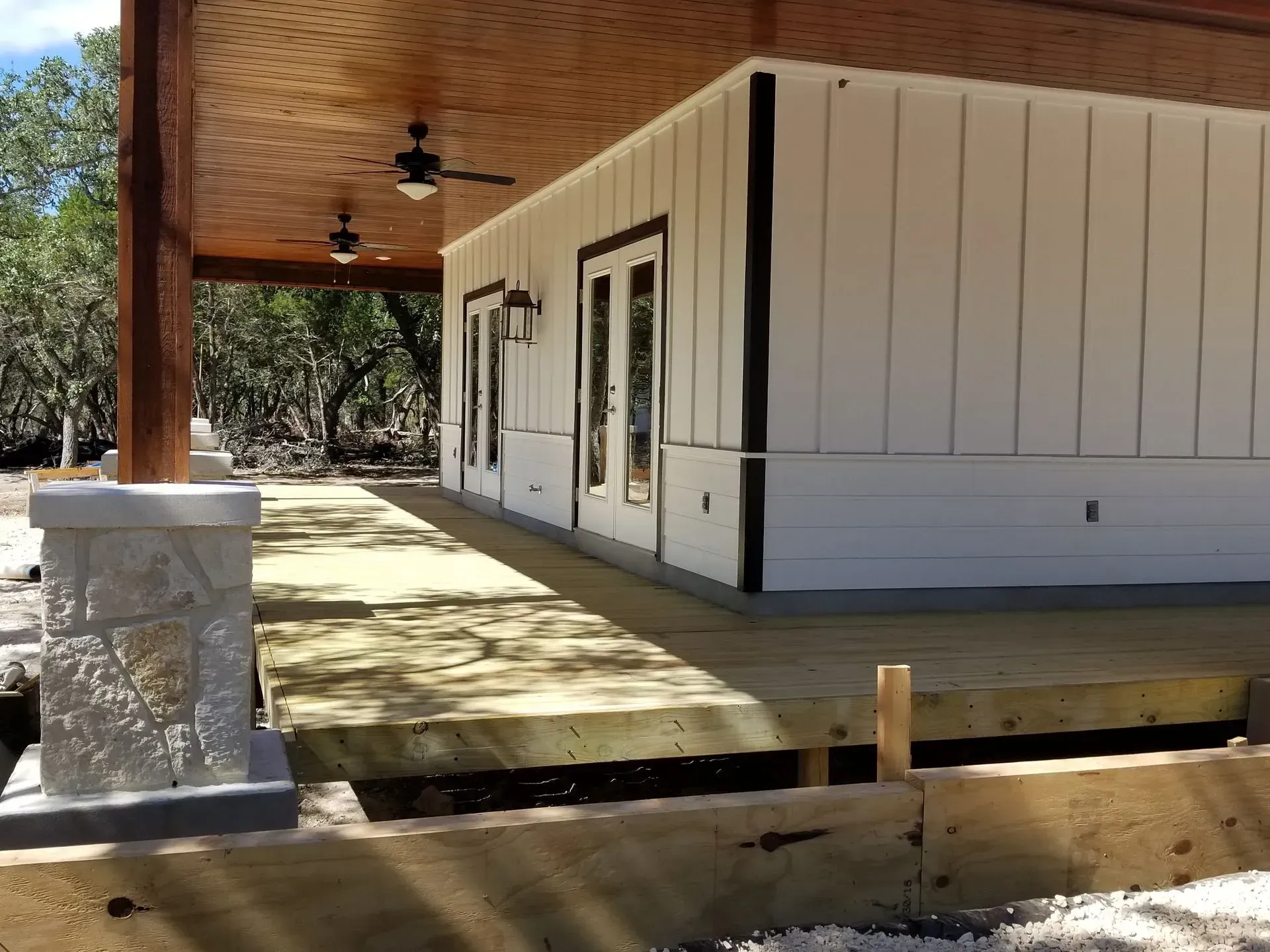 A partially built house with a porch and wooden deck. White siding, black trim. Beige wooden ceiling.