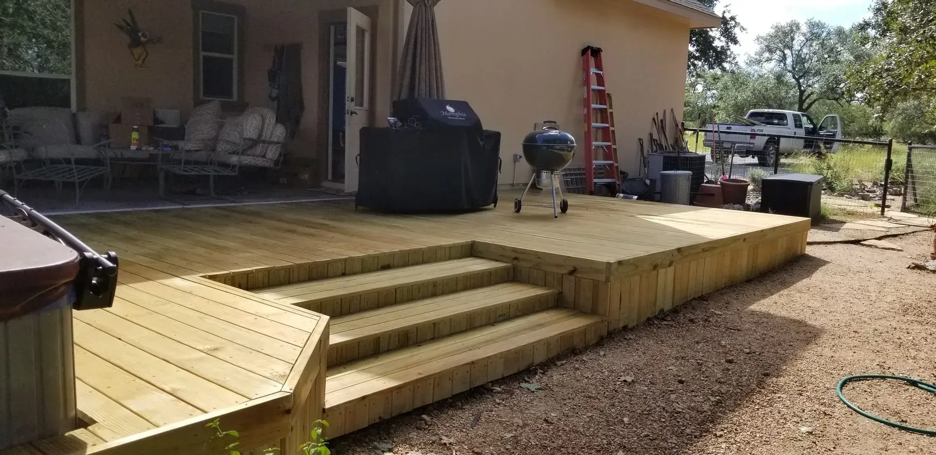 Wooden deck with steps leading to a raised platform. A house is visible, along with a grill and other outdoor items.