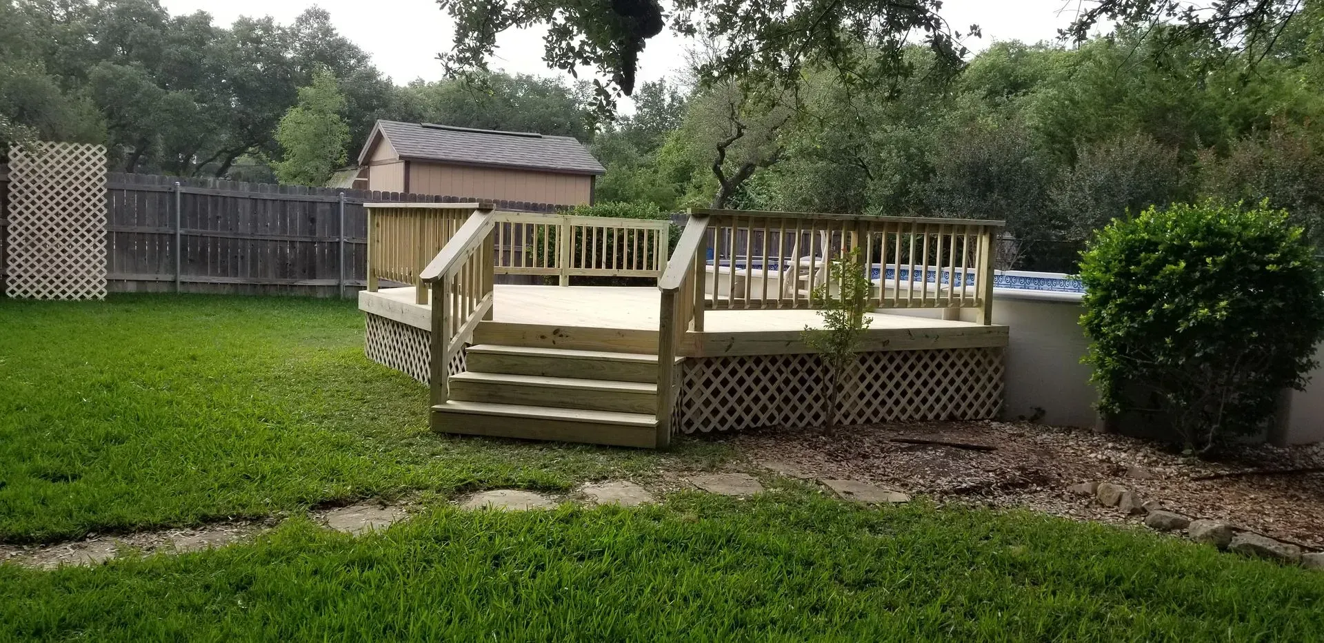 Wooden deck with steps in a backyard. The deck is in front of a shed and a fence. Green grass surrounds it.