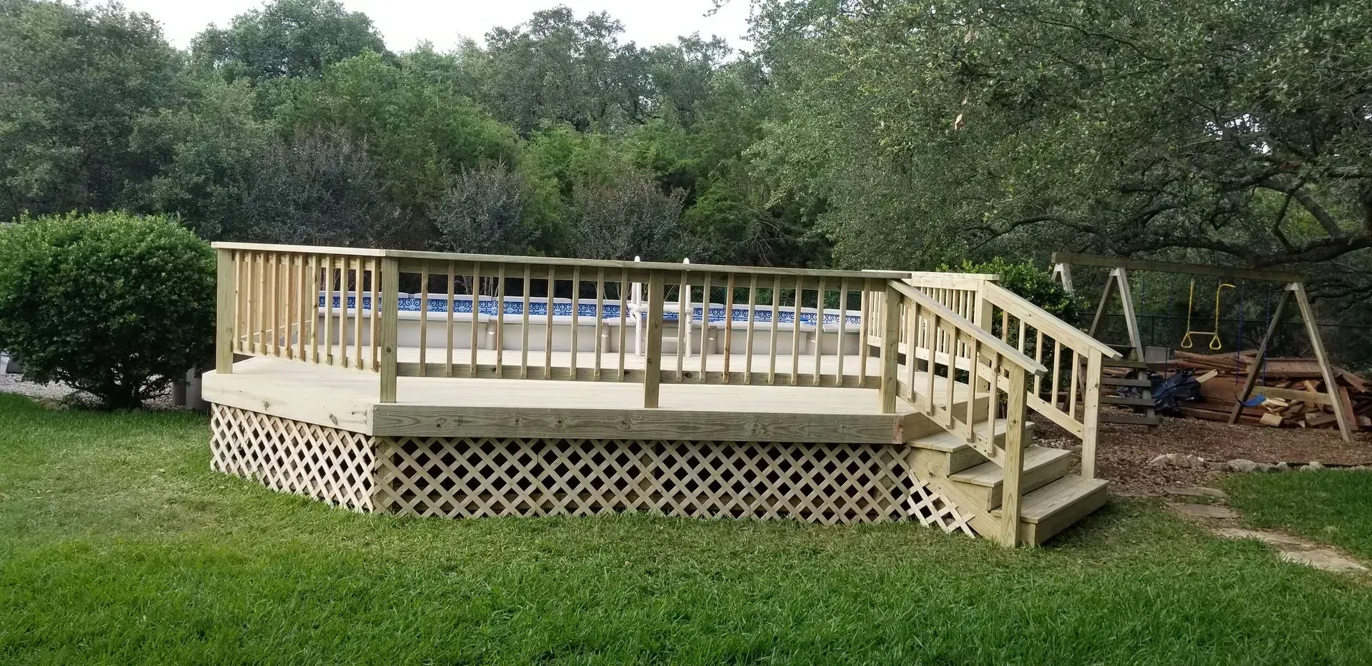 Wooden deck with lattice skirting surrounds an above-ground pool. Stairs lead down to the grassy yard.