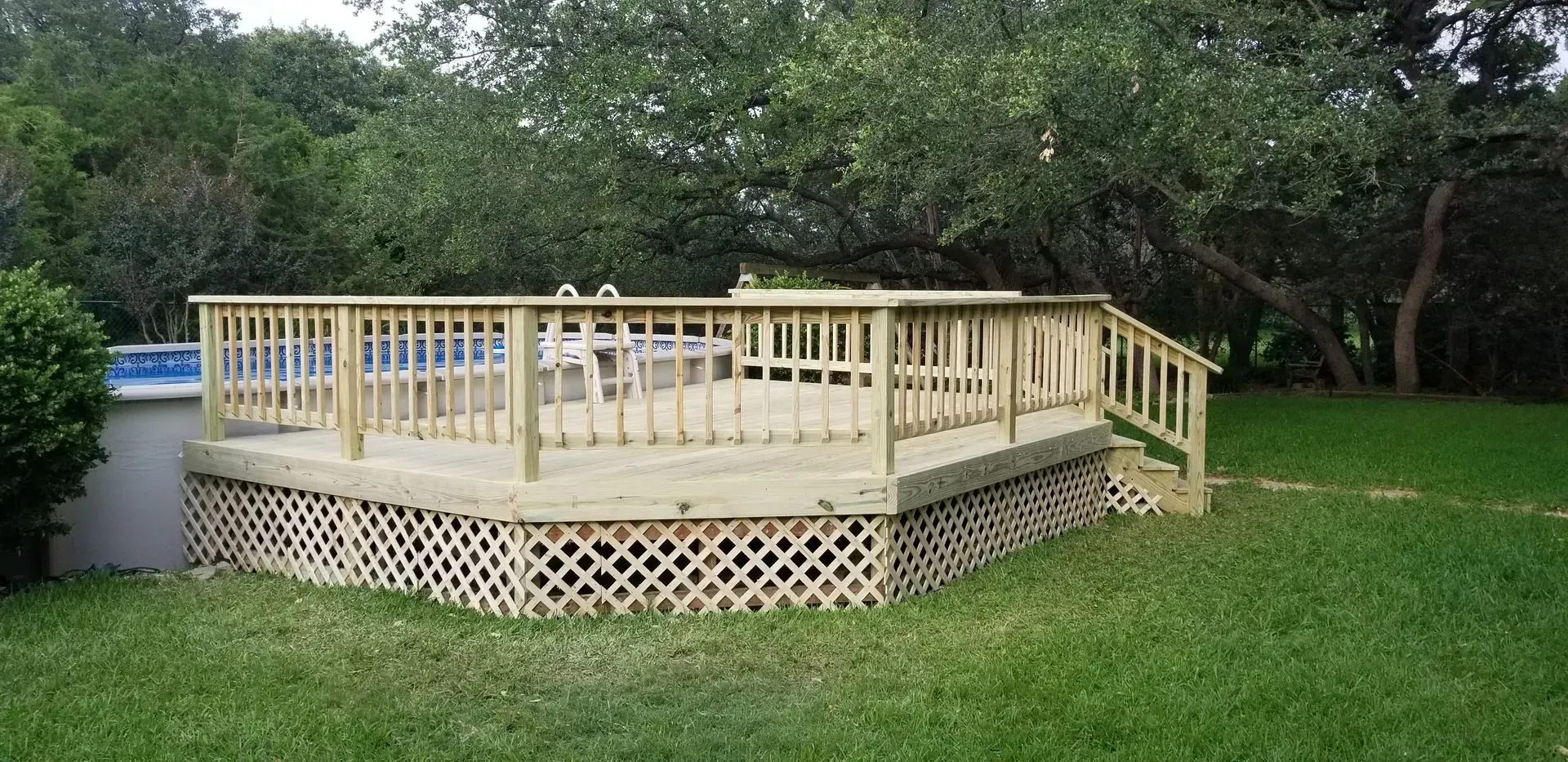 Wooden deck with railing and lattice skirting in a backyard, near a swimming pool.