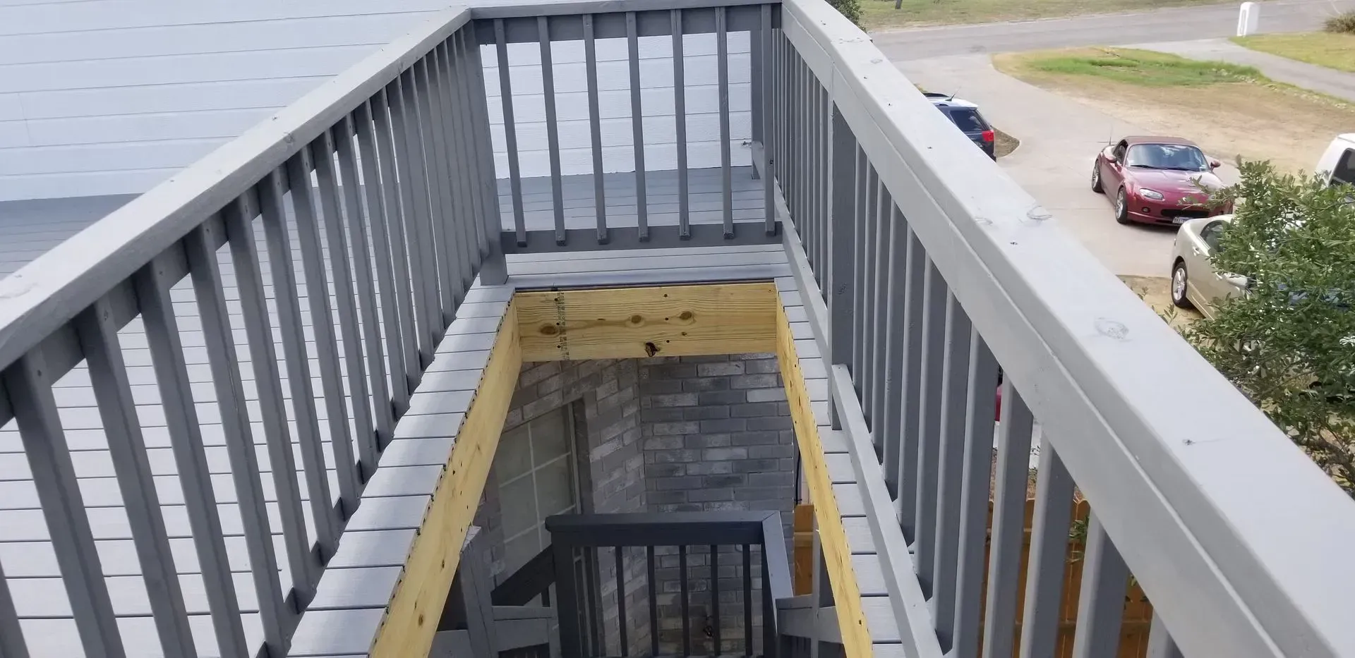 Wooden deck with stairs leading down. Gray railings and a view of parked cars.