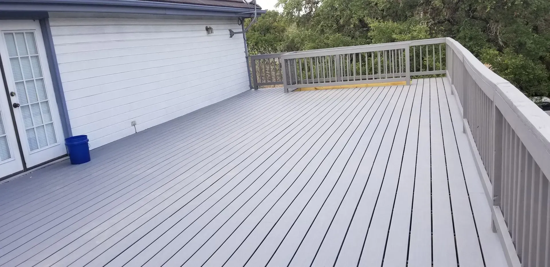 Gray wooden deck with white building and lush greenery in the background.