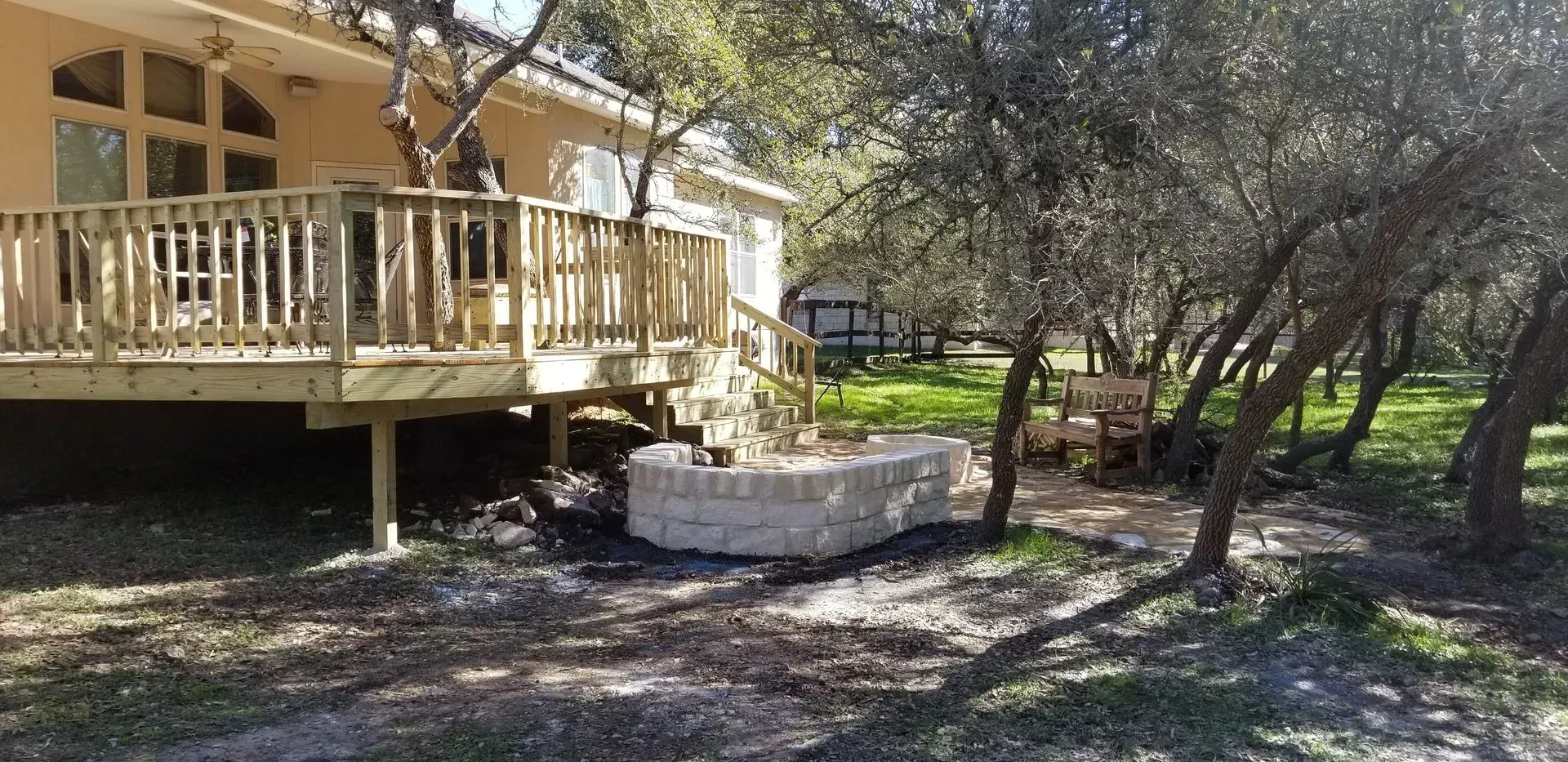Wooden deck on a house with a stone fire pit in a grassy yard under trees.