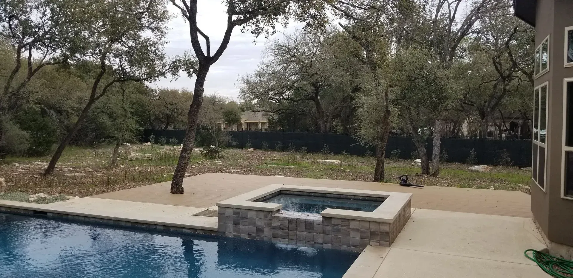 Backyard with pool, hot tub, and trees under a cloudy sky.
