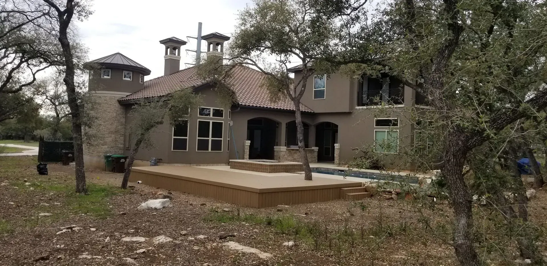 A two-story brown house with a tile roof and a stone tower is surrounded by trees.