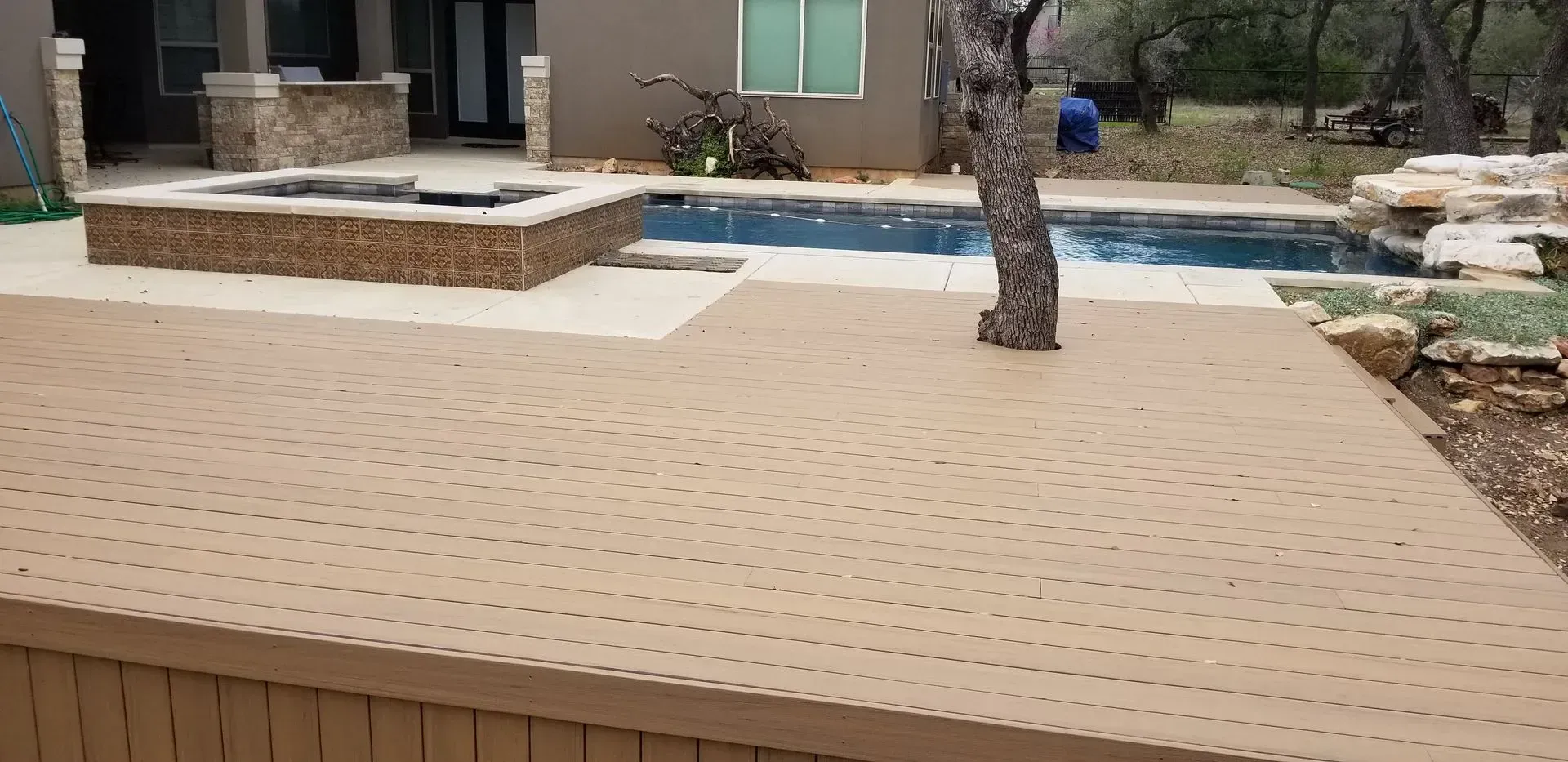 A backyard with a pool, a deck, a hot tub, and a tree.