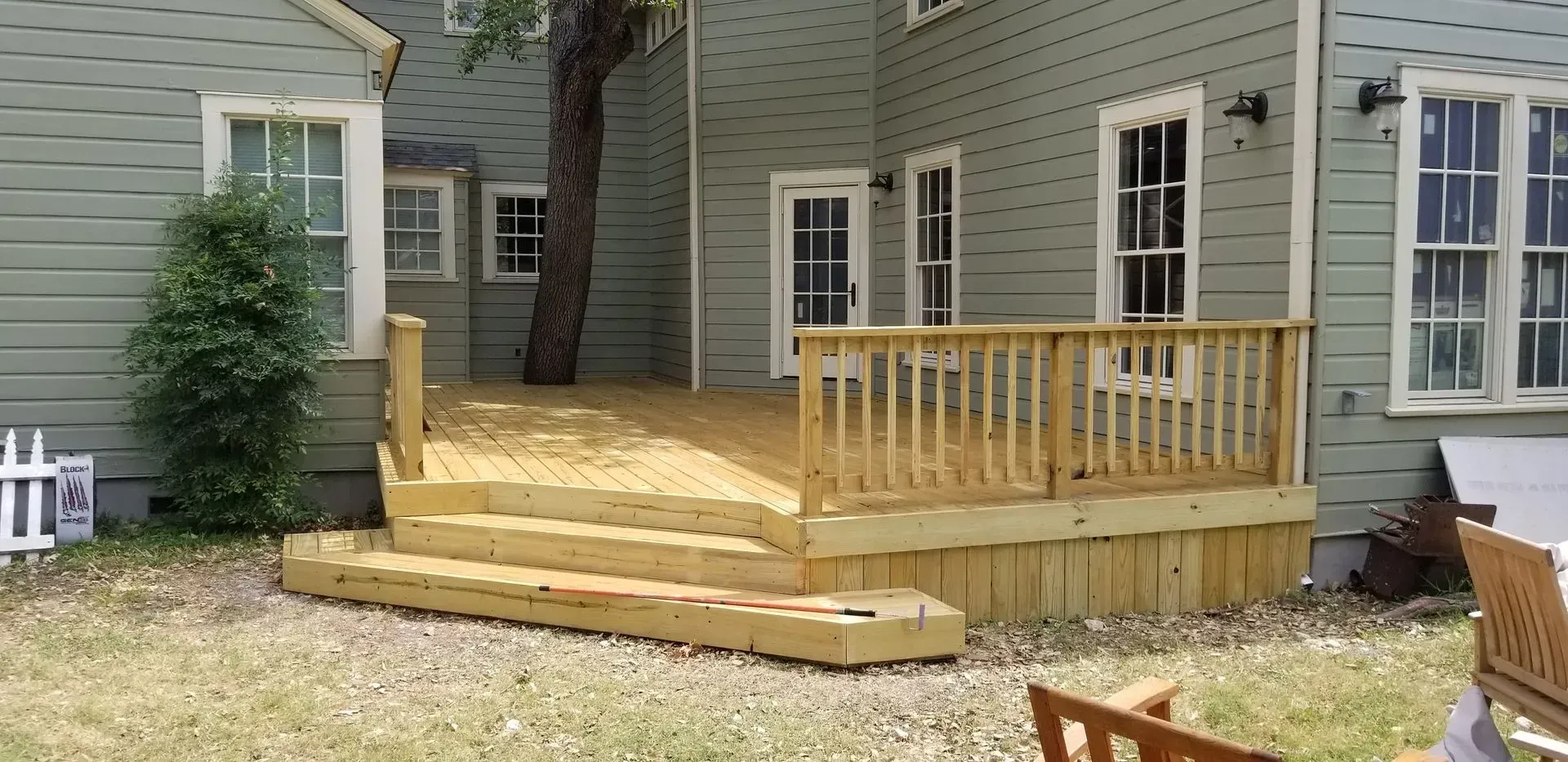 A wooden deck with steps and railing against a green house with a tree growing through it.