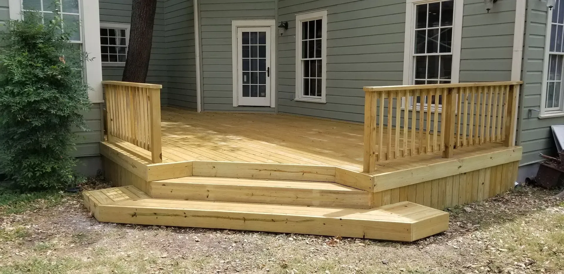 Wooden deck with stairs, railing, and a tree trunk, adjacent to a green house.