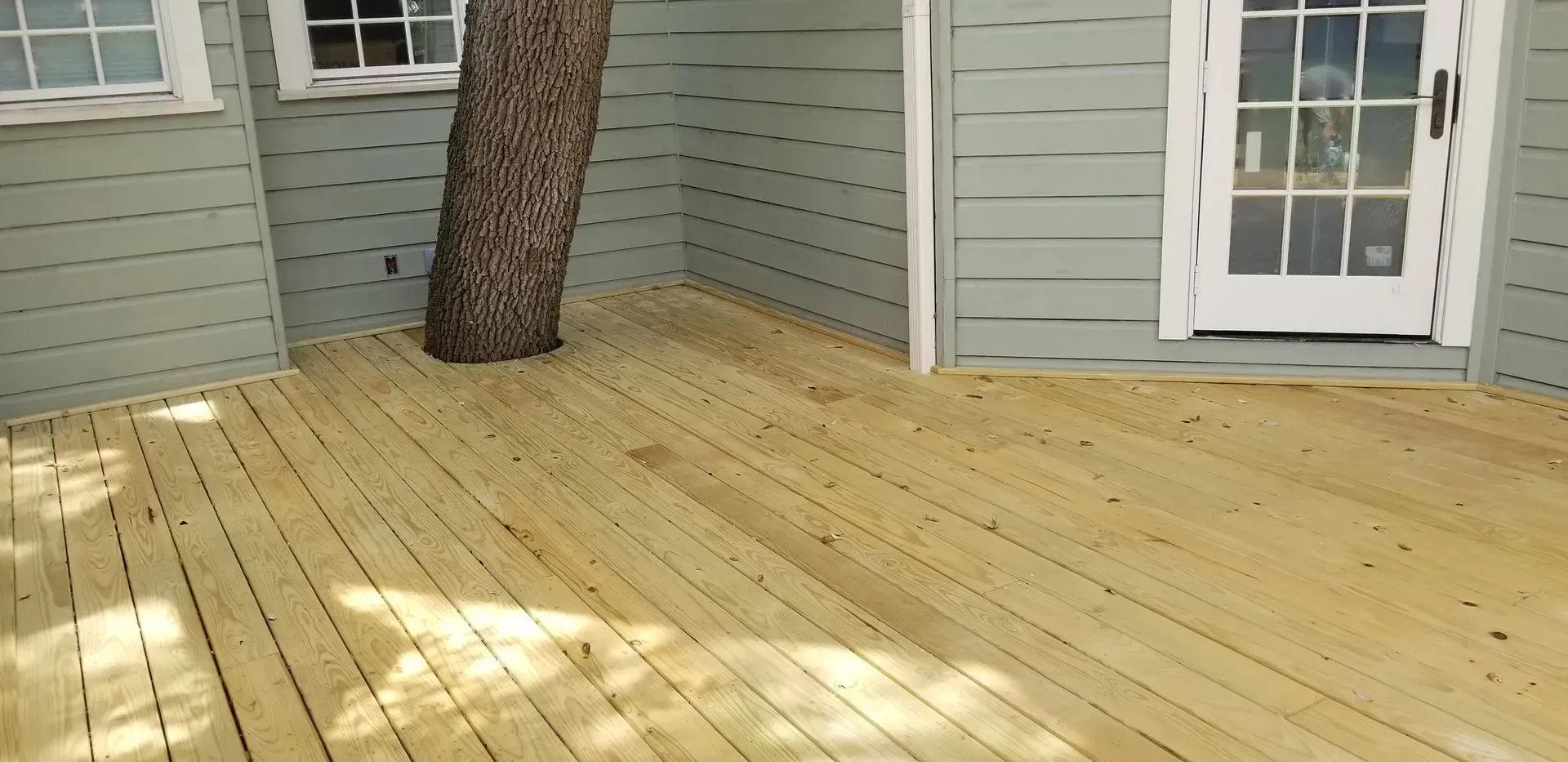 Wooden deck surrounds a tree, next to a light blue house with a white door.