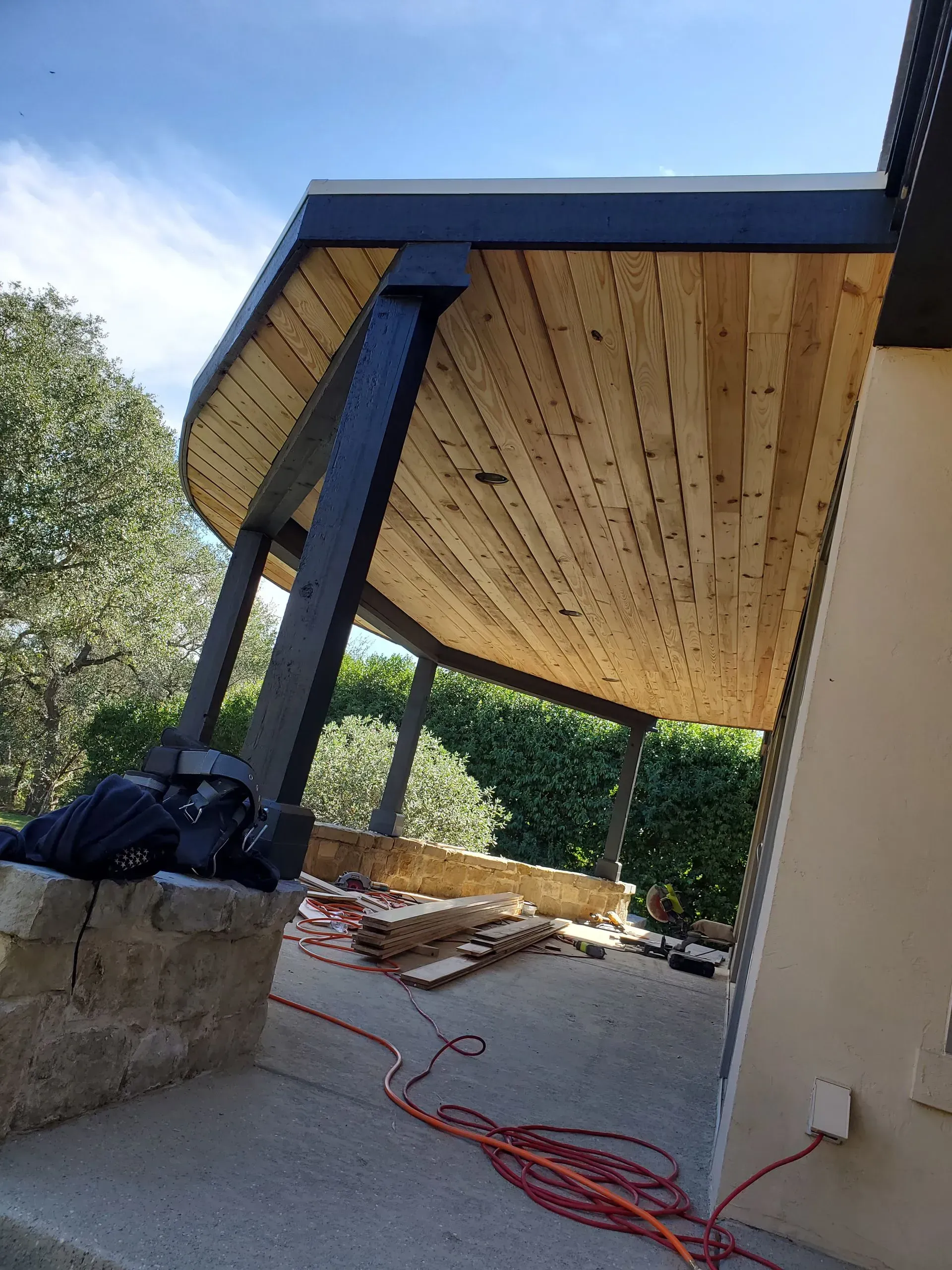 Wooden ceiling of a curved patio with black support beams, a concrete base, and greenery in the background.