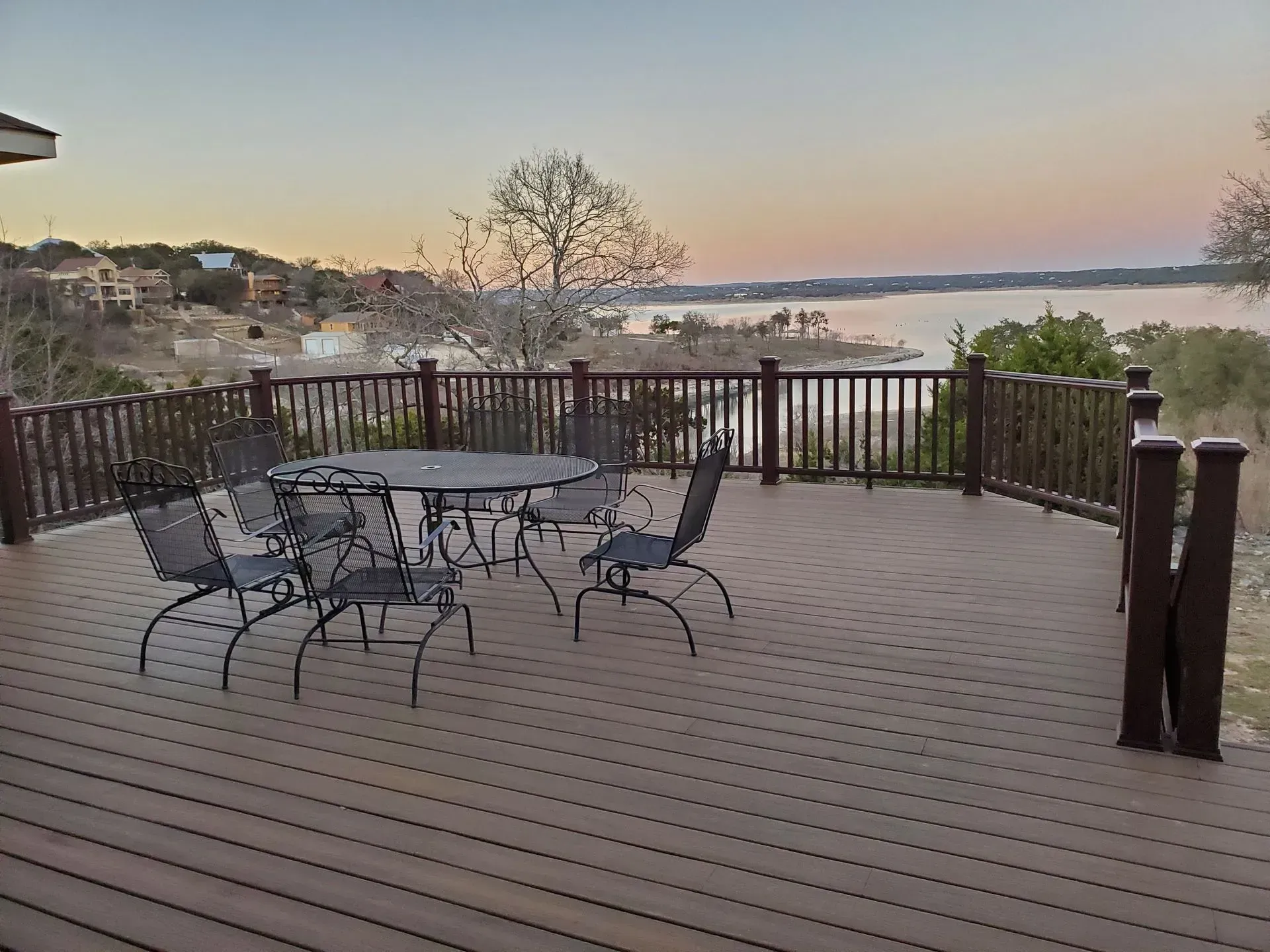Deck overlooking a lake at dusk, with round table and chairs, brown railing.