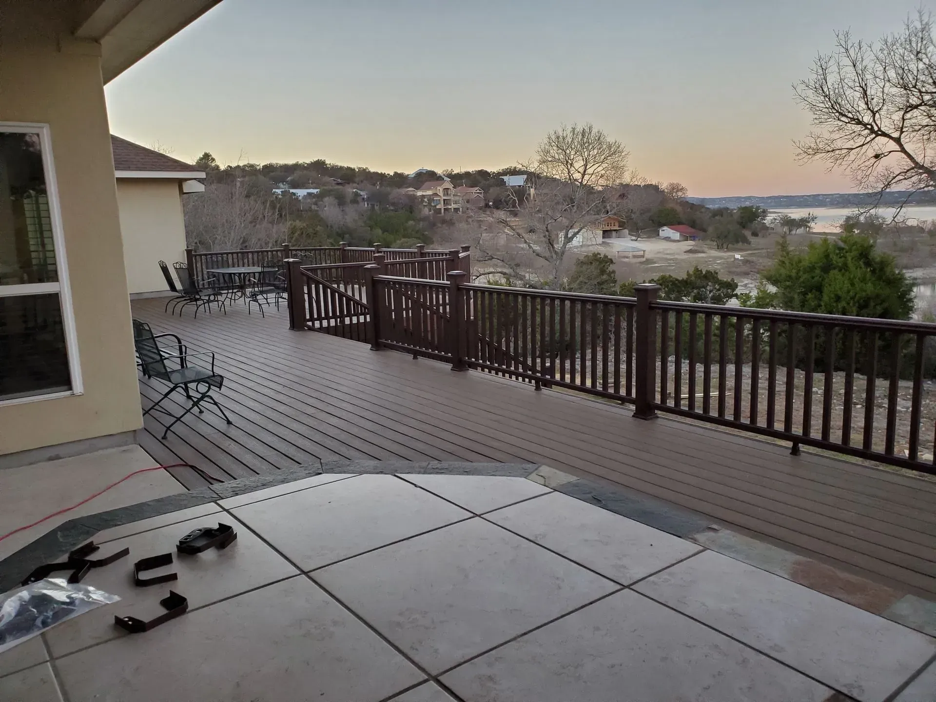 Deck with brown railing overlooks a lake, featuring patio furniture and house.