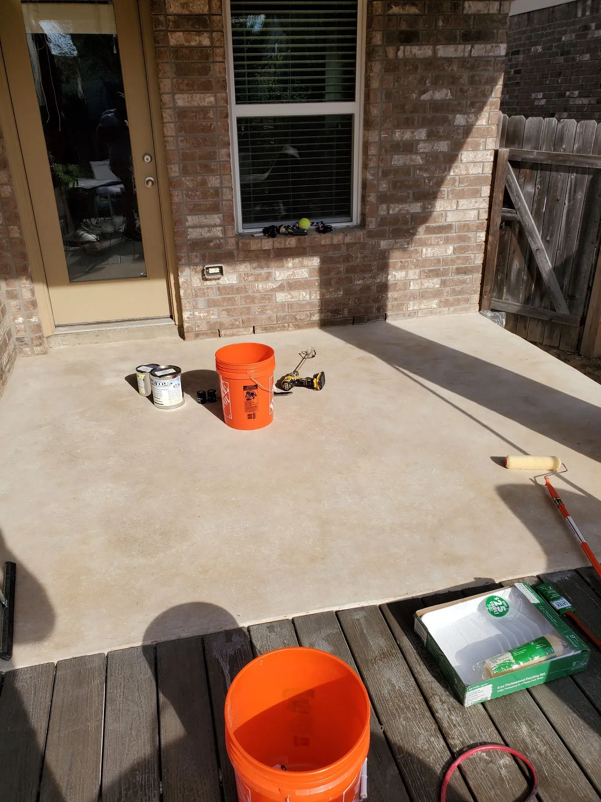 Outdoor patio with tools and an orange bucket on a concrete surface, near a wooden deck and brick wall.
