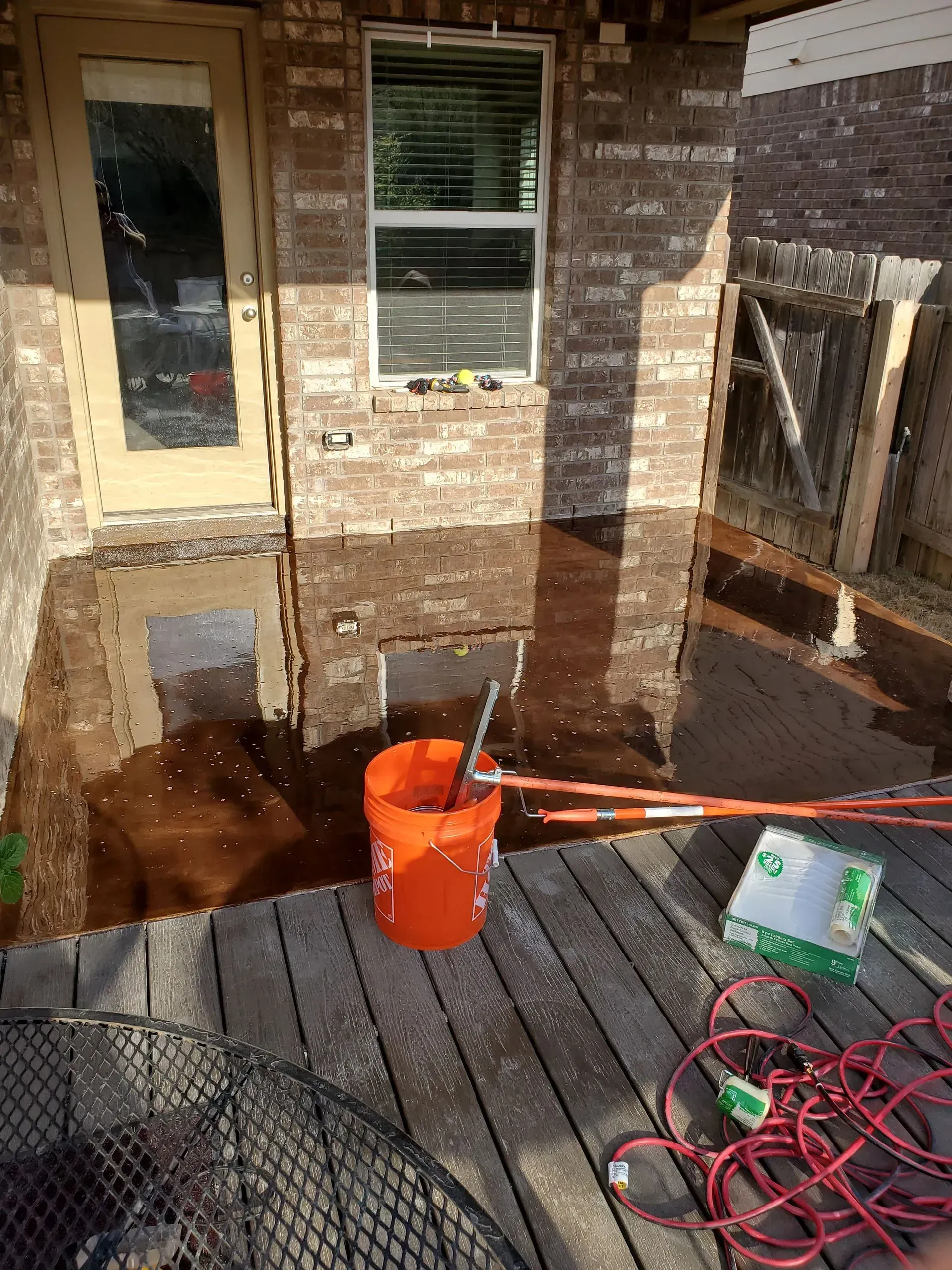 Backyard porch with pooled water. Brick walls, door, window, orange bucket, tools, and deck.