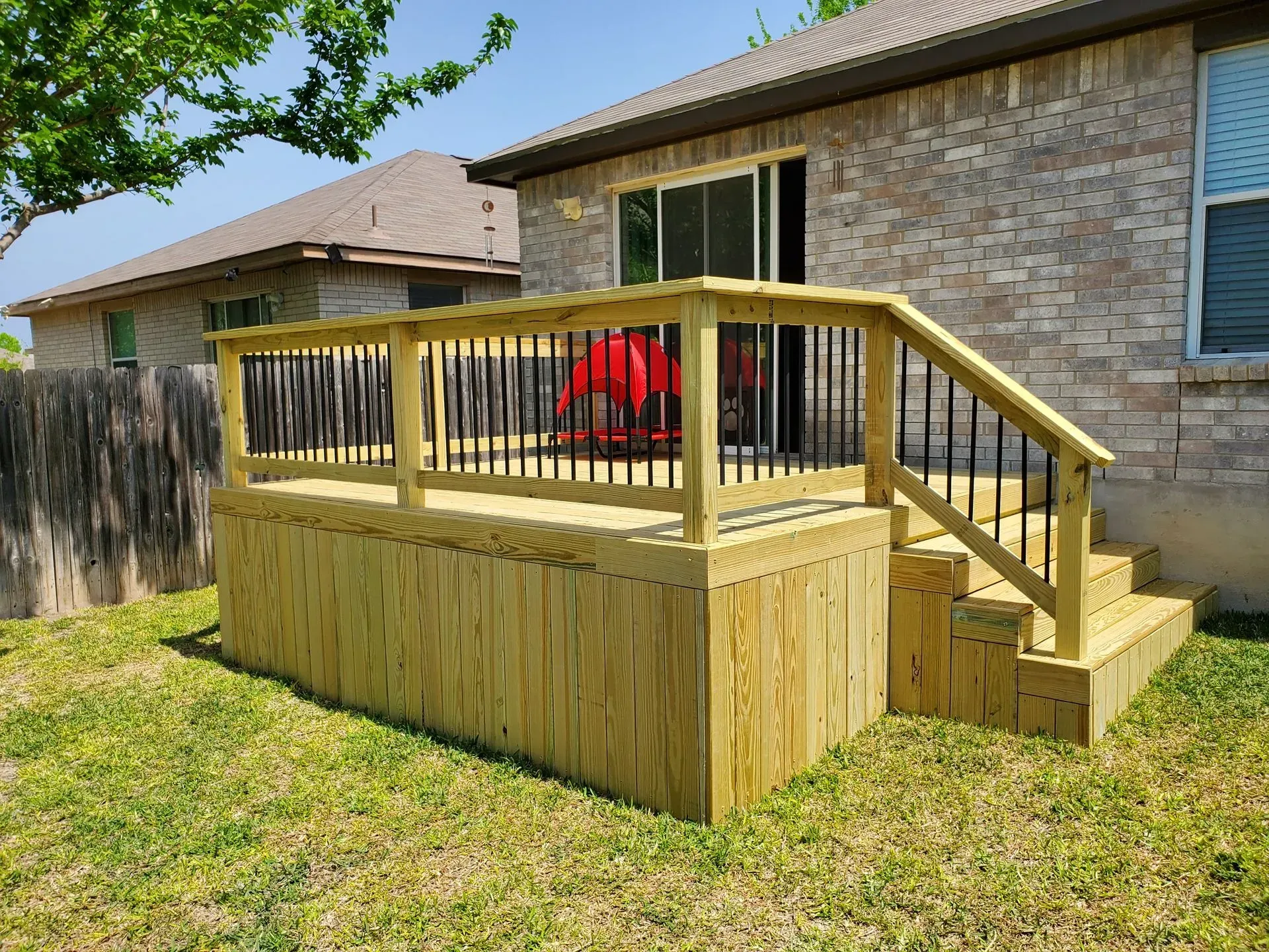 Wooden deck with black railings and steps leading to a sliding glass door. Brick house in background.