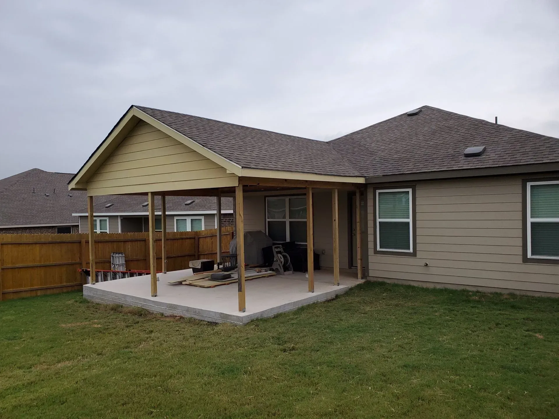 Backyard patio with a covered seating area attached to a light brown house with green grass.