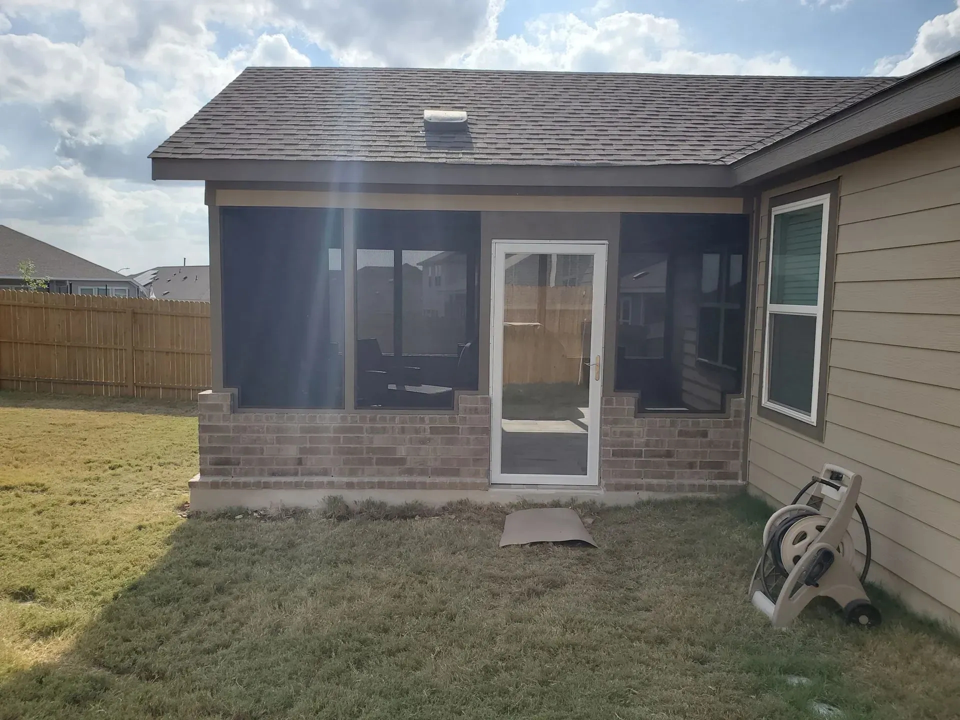 Screened-in porch with brick base and brown roof, adjacent to a beige house, in a grassy backyard.