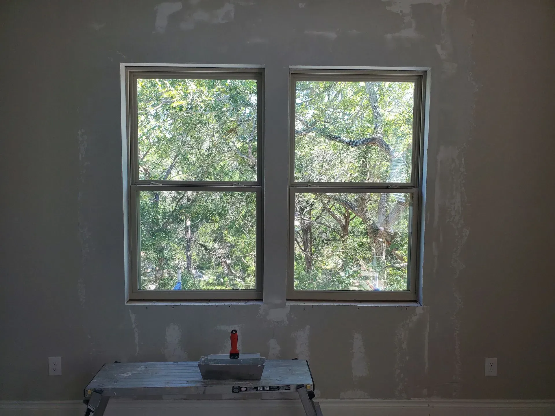 Two vertical windows in a room, looking out onto trees. A roller rests on a small work stand below the windows.
