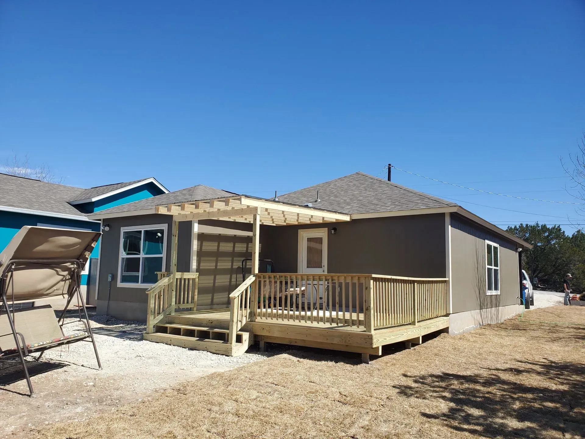 Back of a house with a wooden deck and pergola. Light brown siding, clear blue sky, gravel yard.