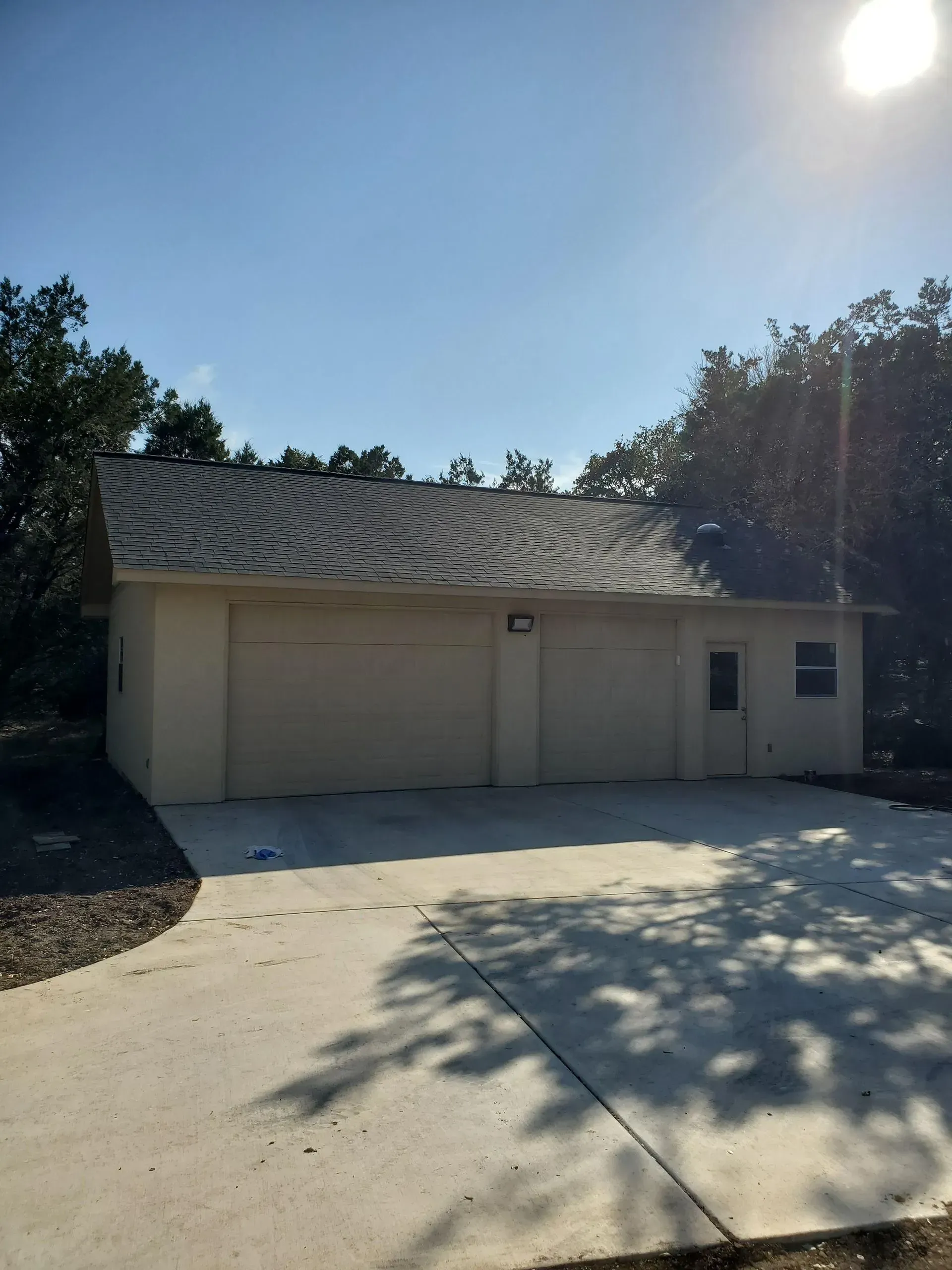 A light beige two-car garage with a gray shingled roof, set against a backdrop of trees, with a concrete driveway.