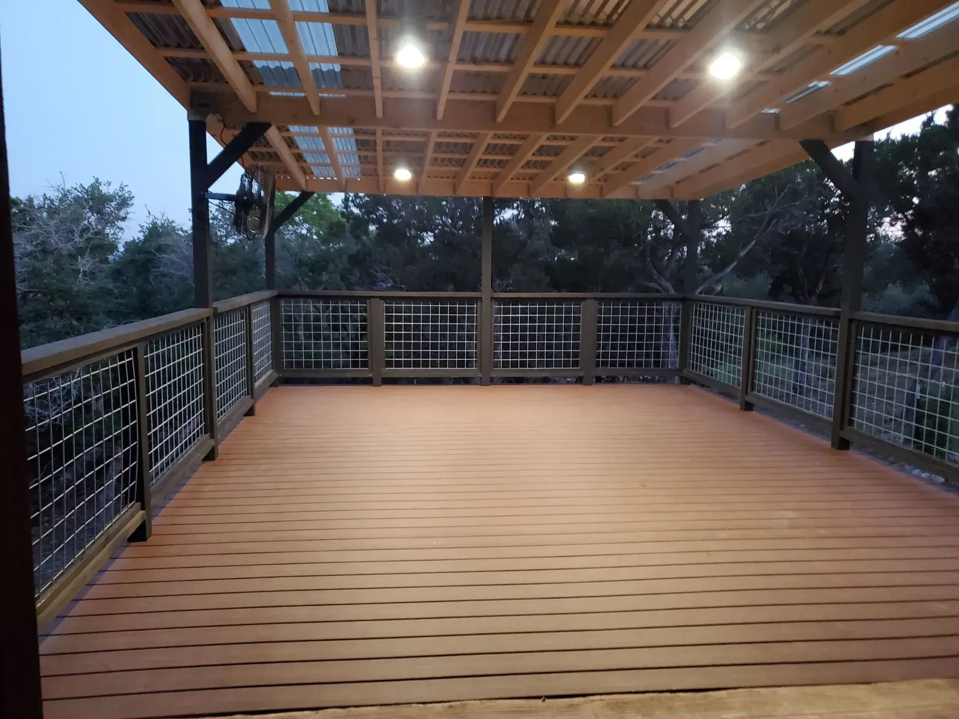 Wooden deck with railing, covered by a wood and corrugated roof, overlooking a wooded area at dusk.