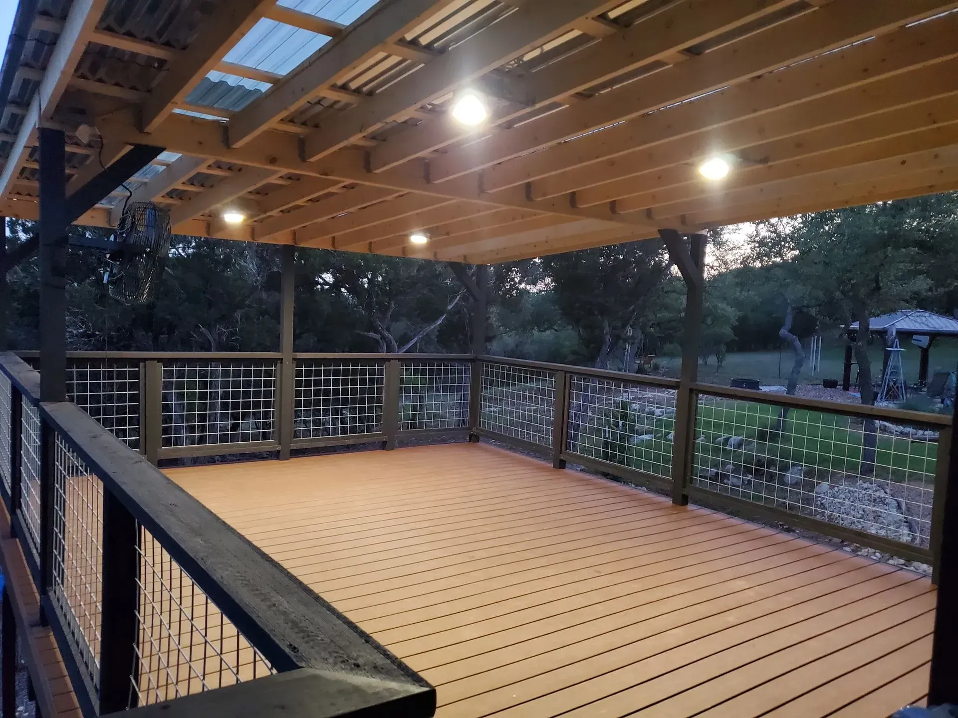 Wooden deck with overhead lighting, overlooking a wooded area. Decorative railing.