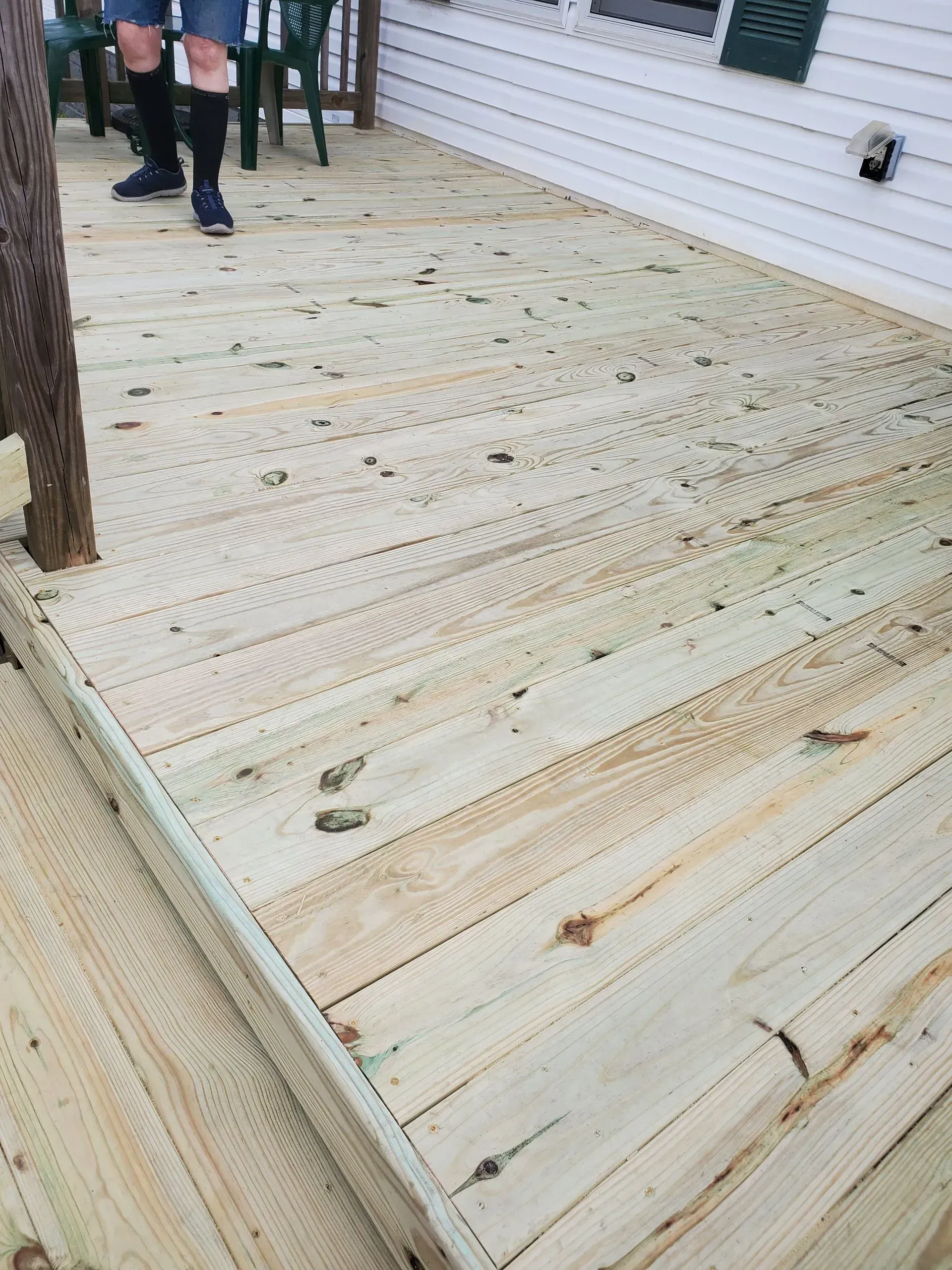Wooden deck with visible tire tracks. Someone stands near green lawn chairs, white siding in background.