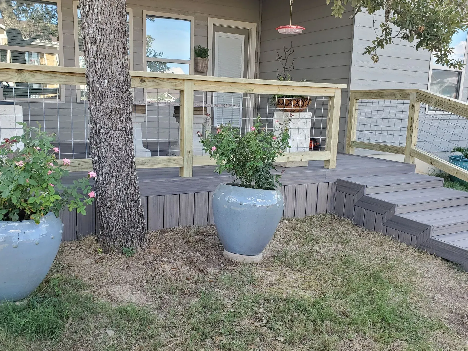 Front porch with gray composite decking, wooden railing, two potted plants, and a small tree.