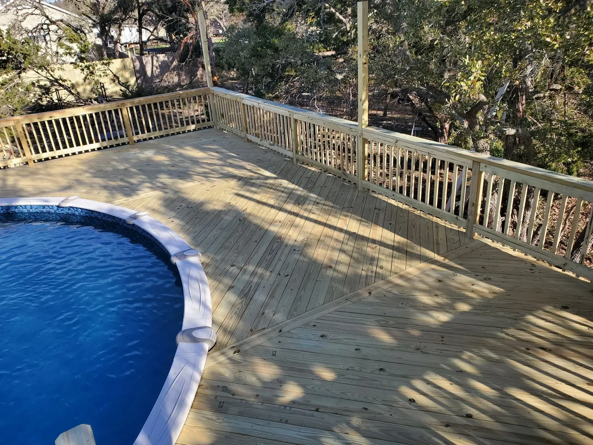 Wooden deck surrounding a blue swimming pool, with a wooden railing and trees in the background.