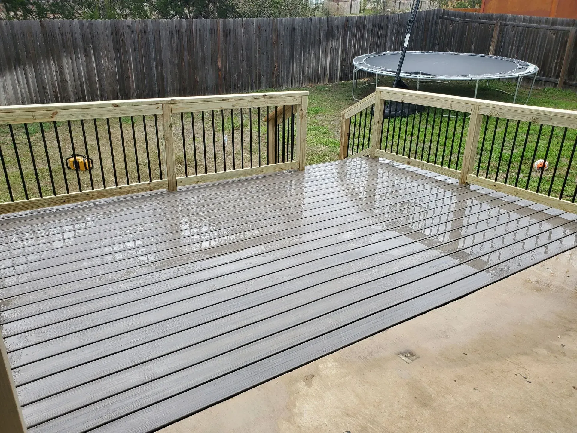 Wet gray composite deck with wooden railings, overlooking a backyard with a trampoline and fence.
