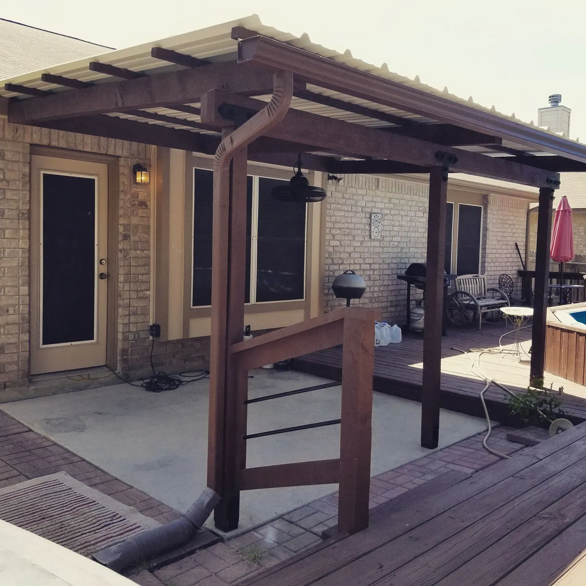 Wooden patio cover with metal roof over concrete patio, brick building background.