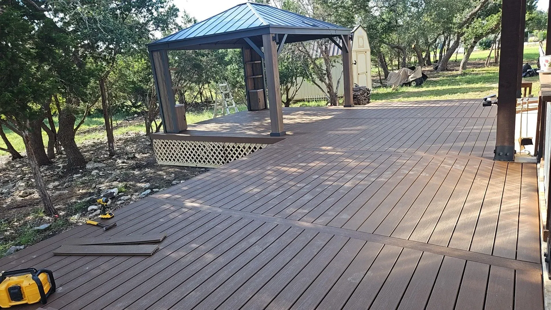 Wooden deck with gazebo in a wooded area; brown boards, green trees, overcast sky.