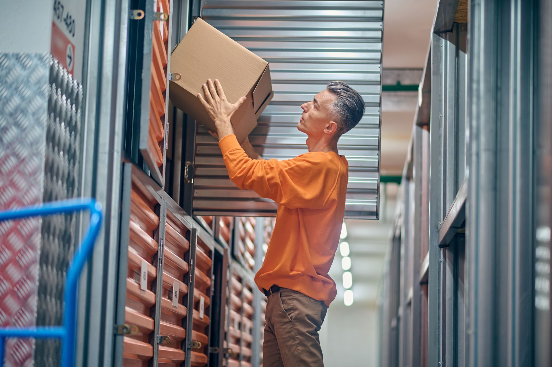 Person placing a cardboard box on a storage shelf. He wears an orange shirt and khaki pants.