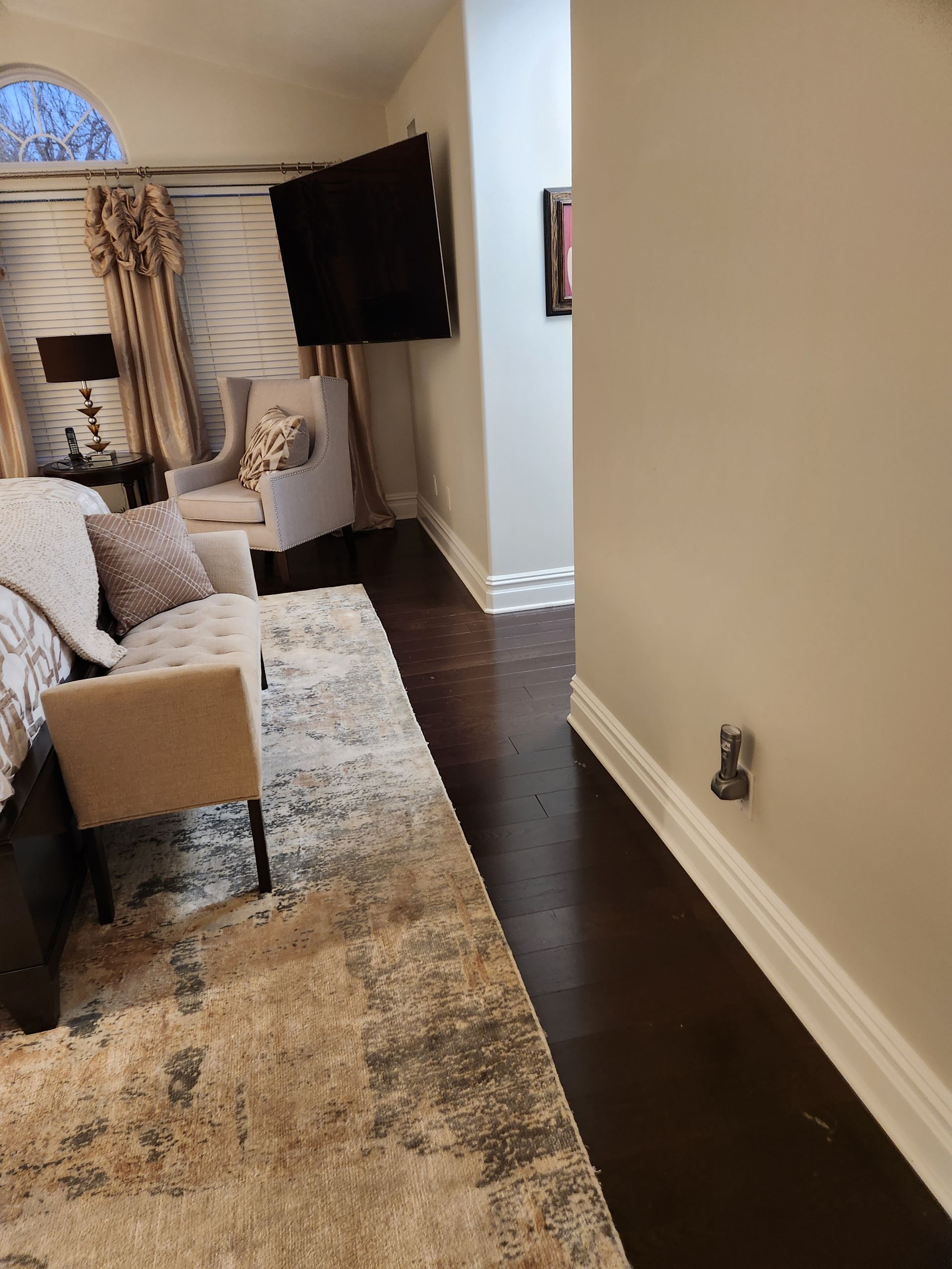 Bedroom with dark wood floors, large rug, mounted TV, seating area with curtains.