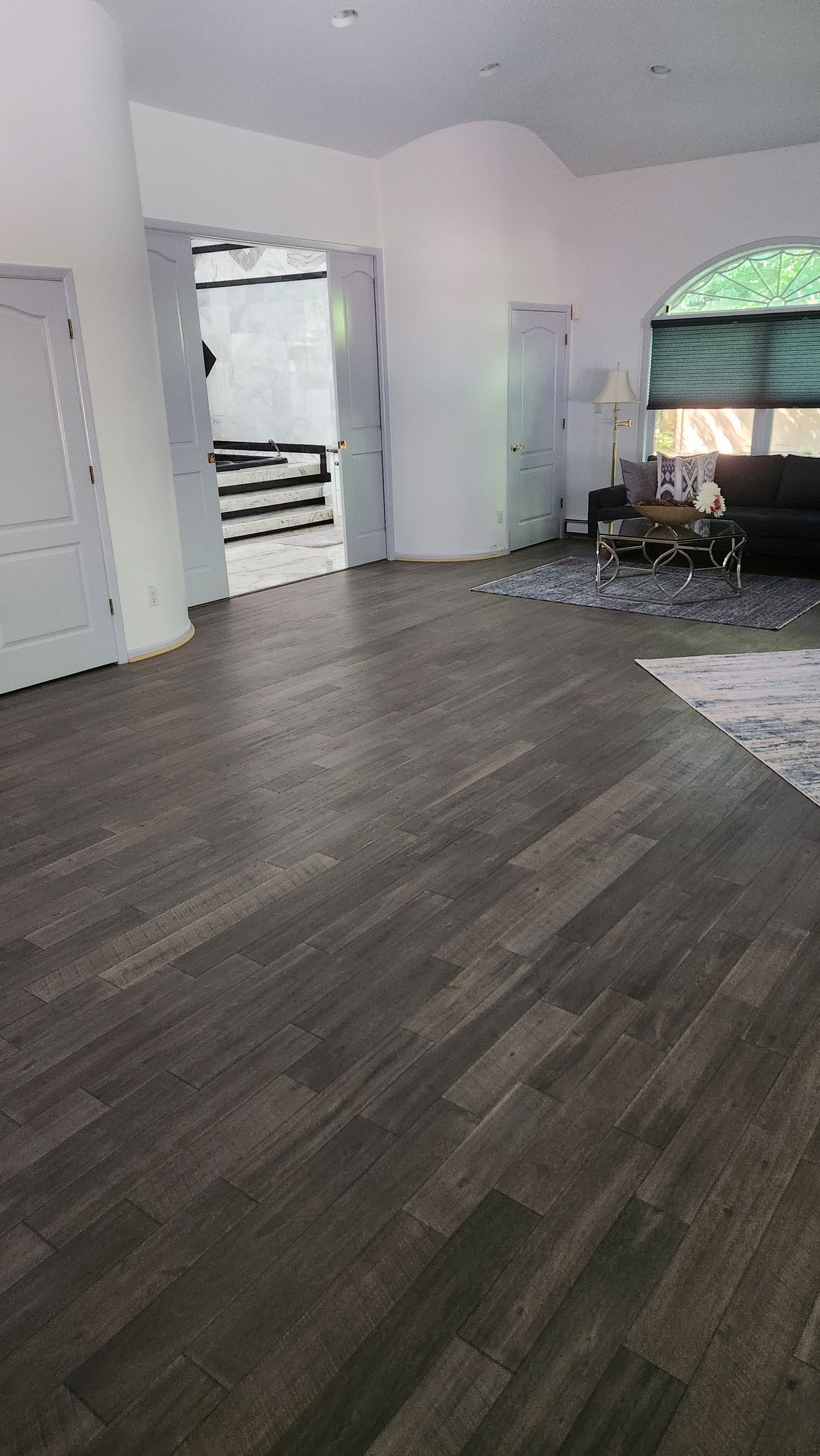 Room with dark wood-look flooring, white walls, and a view into another space. A black sofa sits by a window.